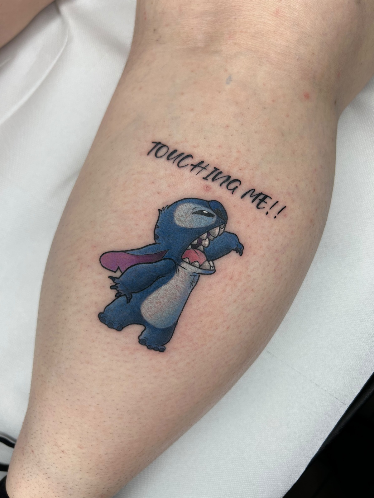 Tatuaggio di Stitch a colori con la scritta "Touching me!", sul polpaccio, parte di un tatuaggio di coppia